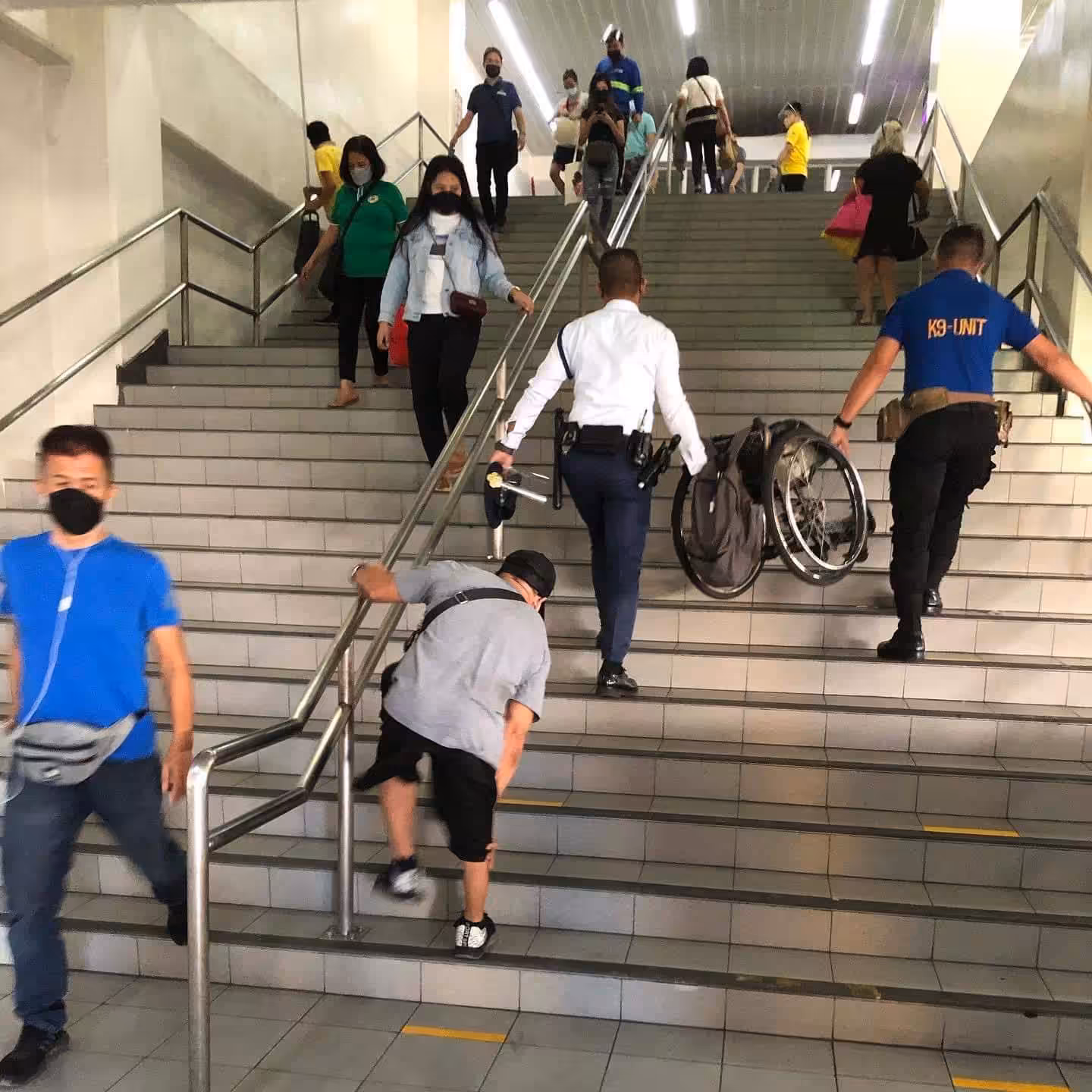 A man with disability climbs the stairs up the train station as 2 employees carry his wheelchair ahead