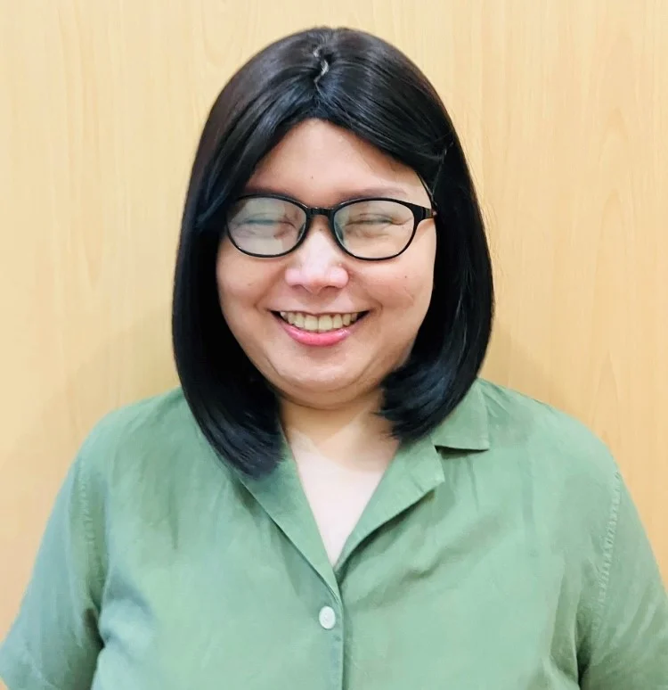 Rhea has straight black hair, glasses, and a green shirt smiling against a wood background.